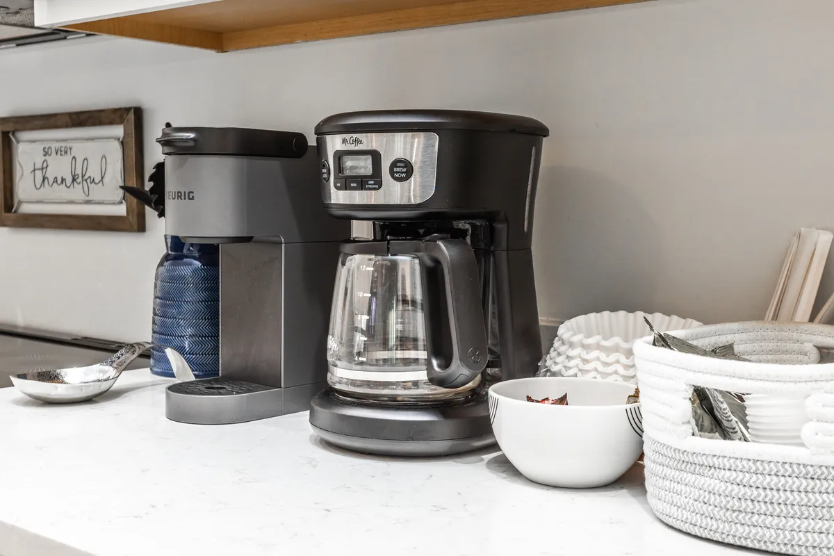 A close-up of a coffee station in the kitchen featuring both a Keurig and a Mr. Coffee machine, with a white bowl and woven basket on the counter. The setup highlights the convenience and warmth of starting the day with a shared cup of coffee.