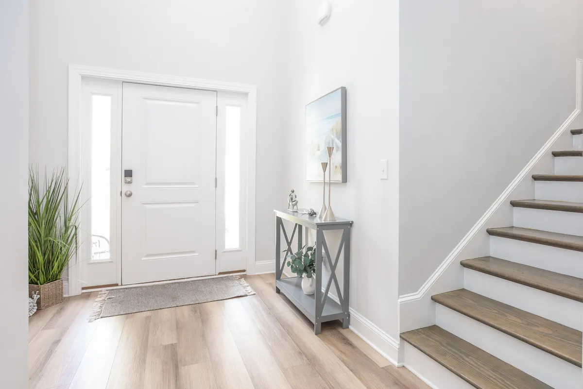 A welcoming entryway with a front door, a console table adorned with decor, and a staircase rising in the background. The space is bright, features an indoor plant, and sets a warm, homey tone as one steps inside.