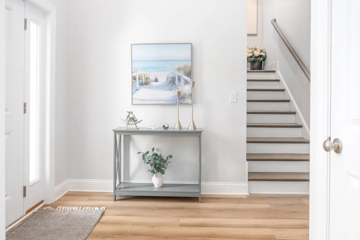 A bright transitional space featuring beach-themed artwork, a console table with decorative pieces, a staircase, and indoor plants. The abundance of natural light and calm palette evoke a serene, coastal vibe.