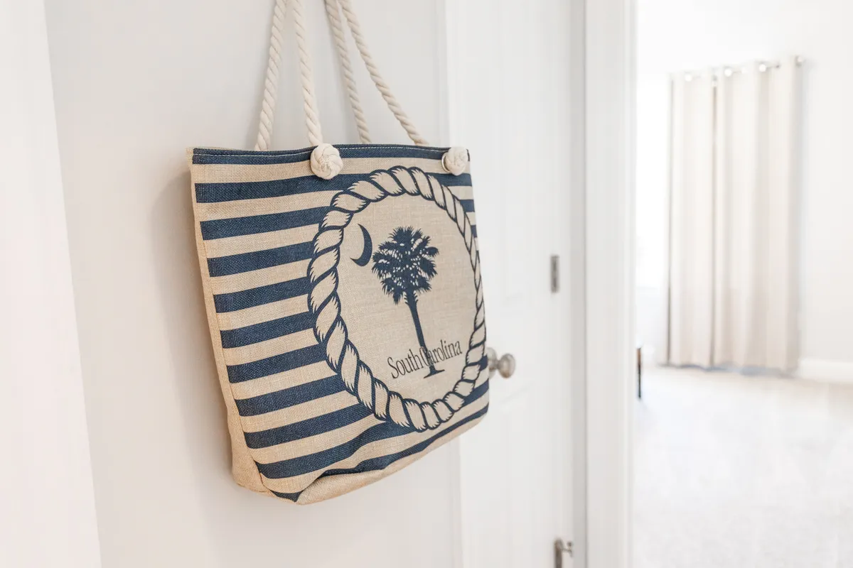 A relaxed entryway scene with a striped beach bag featuring a palm tree design, set by a white door with soft curtains in the background. The casual, beachy vibe evokes the excitement of setting out for or returning from a sunny day on the sand.