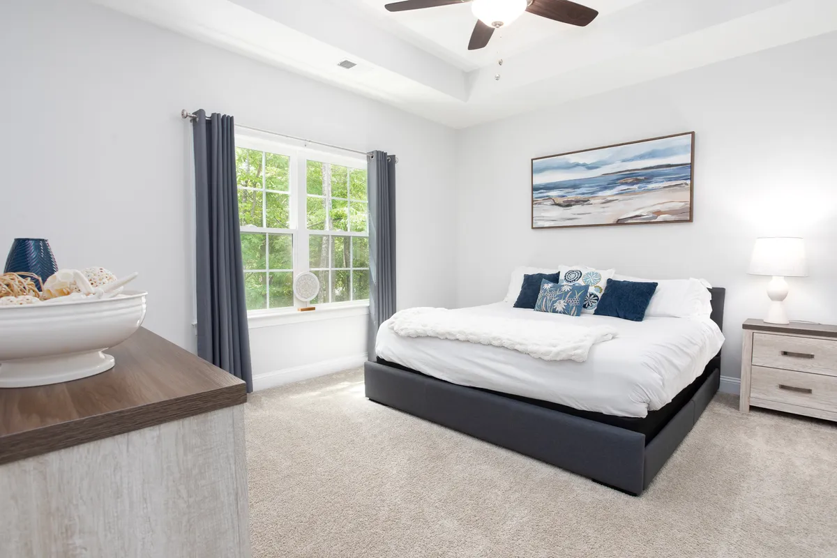 This image features a comfortable bed adorned with decorative pillows, a large window filling the room with natural light, a painting on the wall, and a nightstand with a lamp. The serene mood and soft lighting create a restful, inviting bedroom atmosphere.