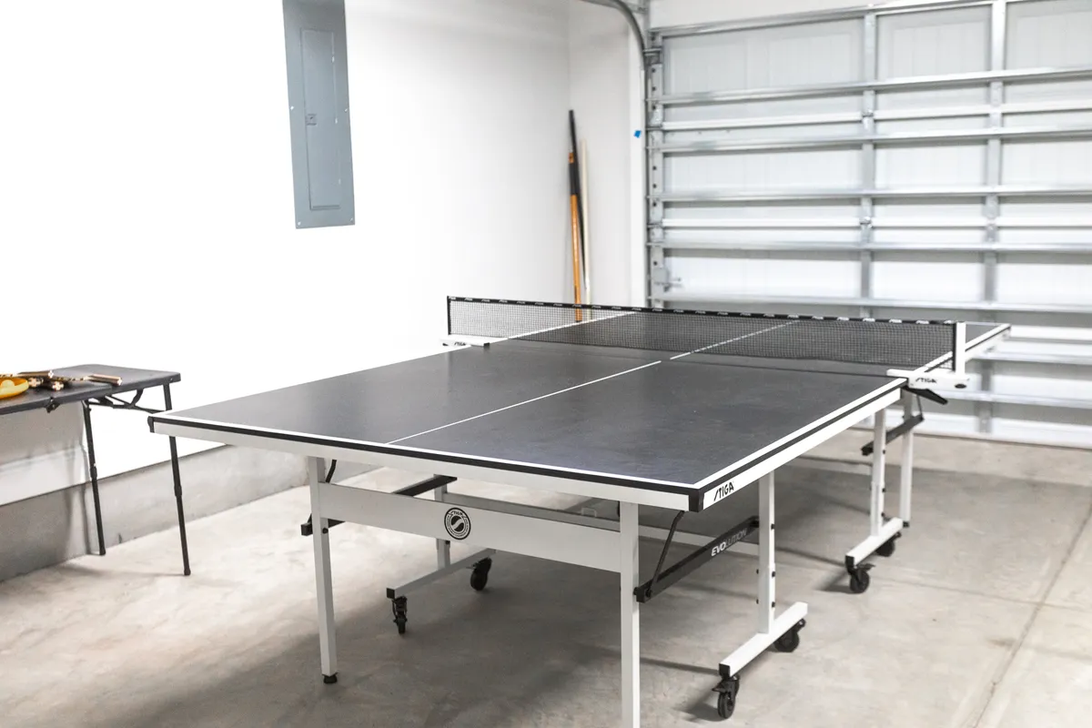 A dedicated game area with a central ping pong table, casual folding table nearby, and visible garage door. The space is clearly designed for play and socializing, with open layout and relaxed atmosphere.