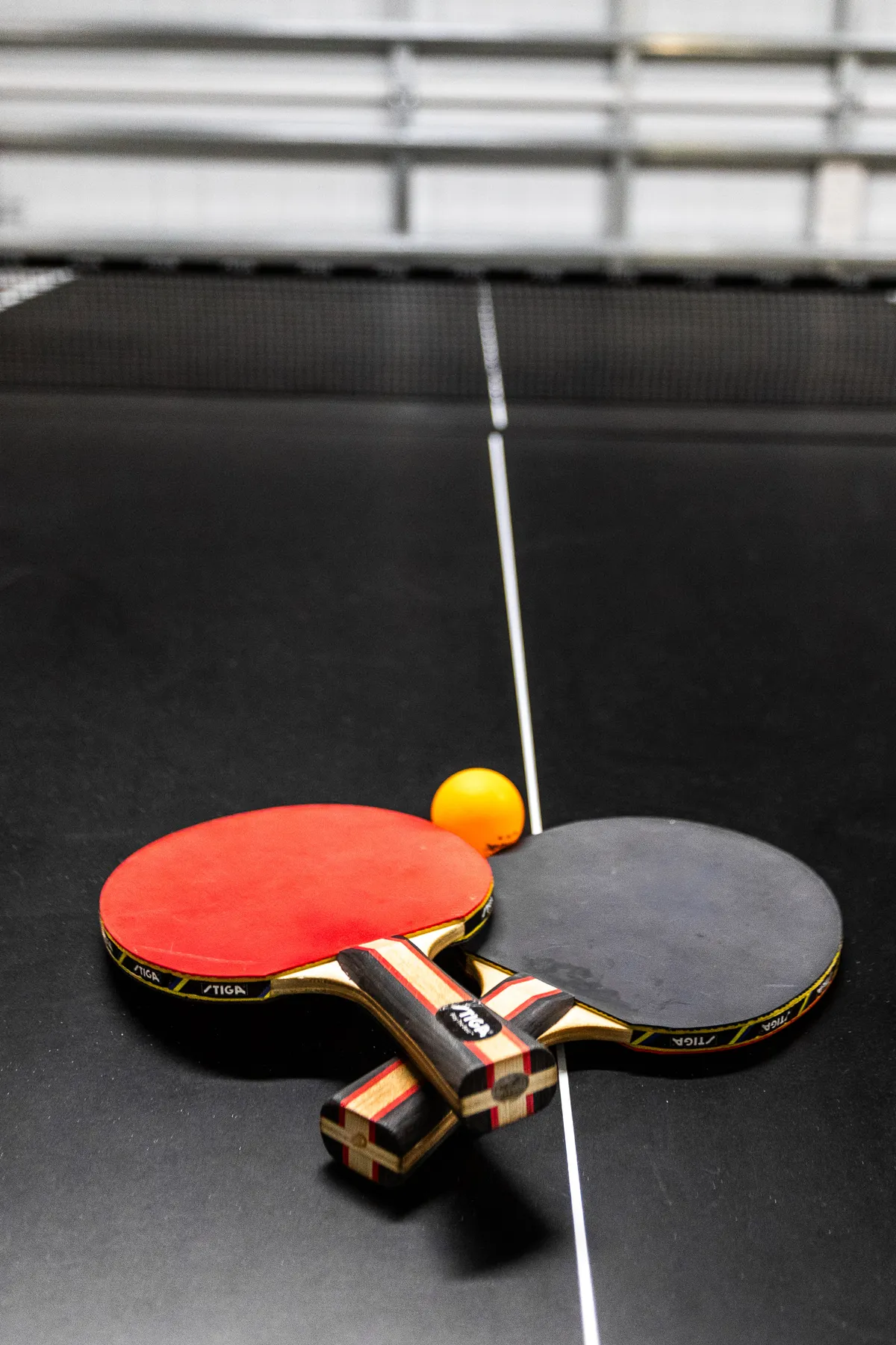 Close-up of two ping pong paddles (red and black) and an orange ping pong ball on a table surface, capturing the anticipation and excitement of a game about to start.
