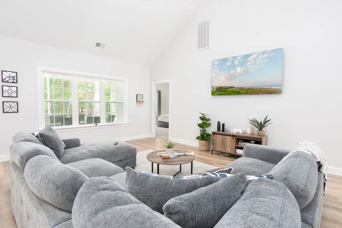 A spacious, light-filled living room featuring a comfortable sectional sofa, large windows streaming in natural light, a television mounted on the wall, a modern coffee table, and lush indoor plants. The setting exudes warmth and is clearly intended for group relaxation, movie nights, and social events.