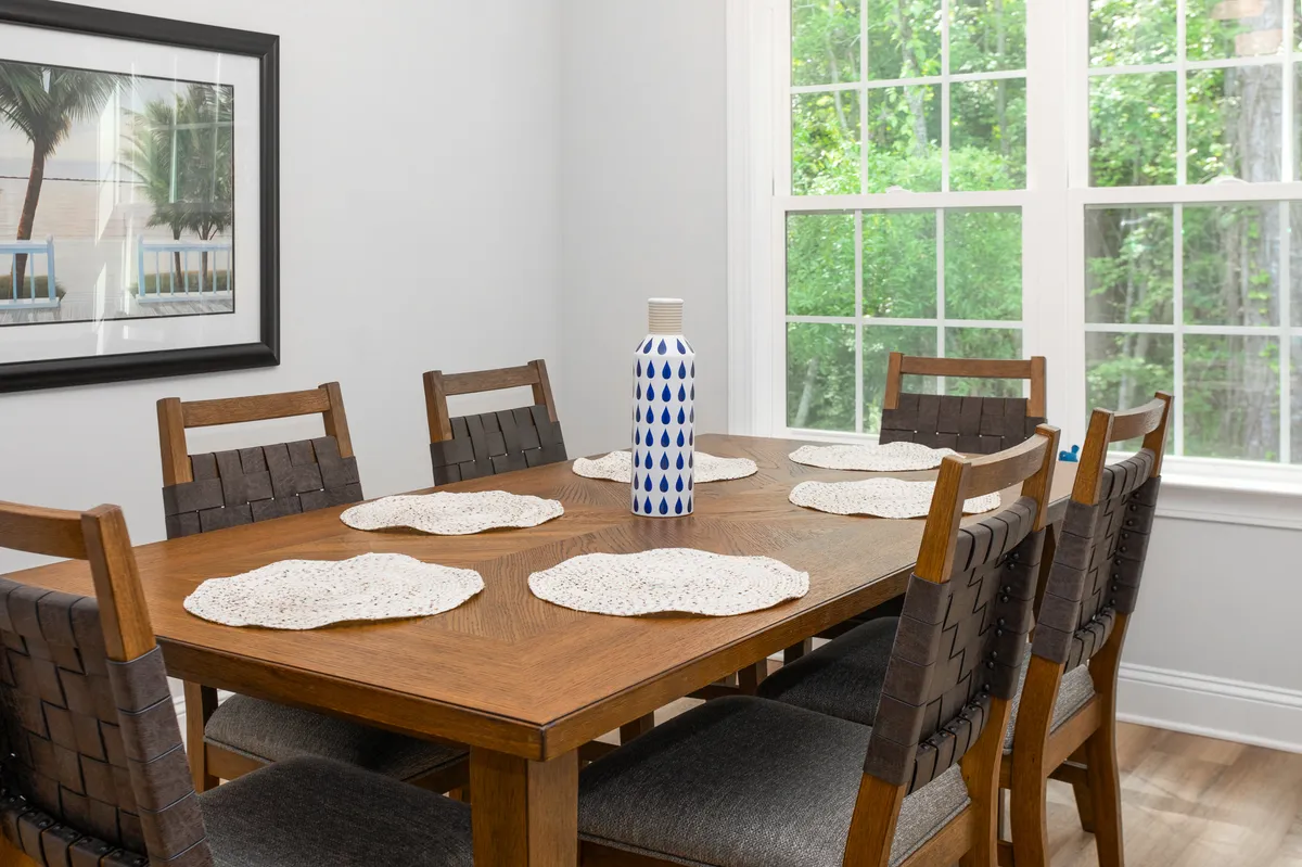 A spacious dining area with a large wooden table, woven placemats, a decorative vase, and framed artwork on the wall. The setting is warm and welcoming, ideal for meals and conversation.