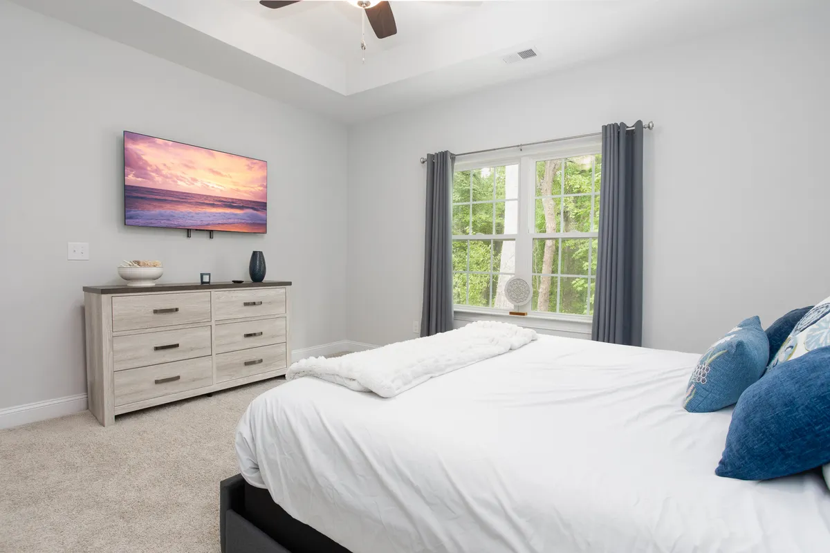 A cozy bedroom with a bed dressed in white bedding, a TV mounted on the wall, a dresser with decorative items, and a window offering a view of greenery outside. The setting exudes a serene and cozy vibe.